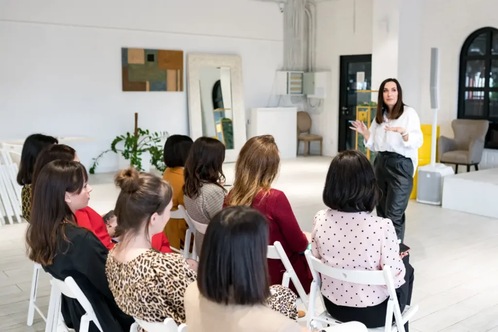A woman stands at the front of a bright, modern room, speaking to a seated audience of attentive women. The room, infused with creative inspiration, features modern decor and mirrors on the walls.